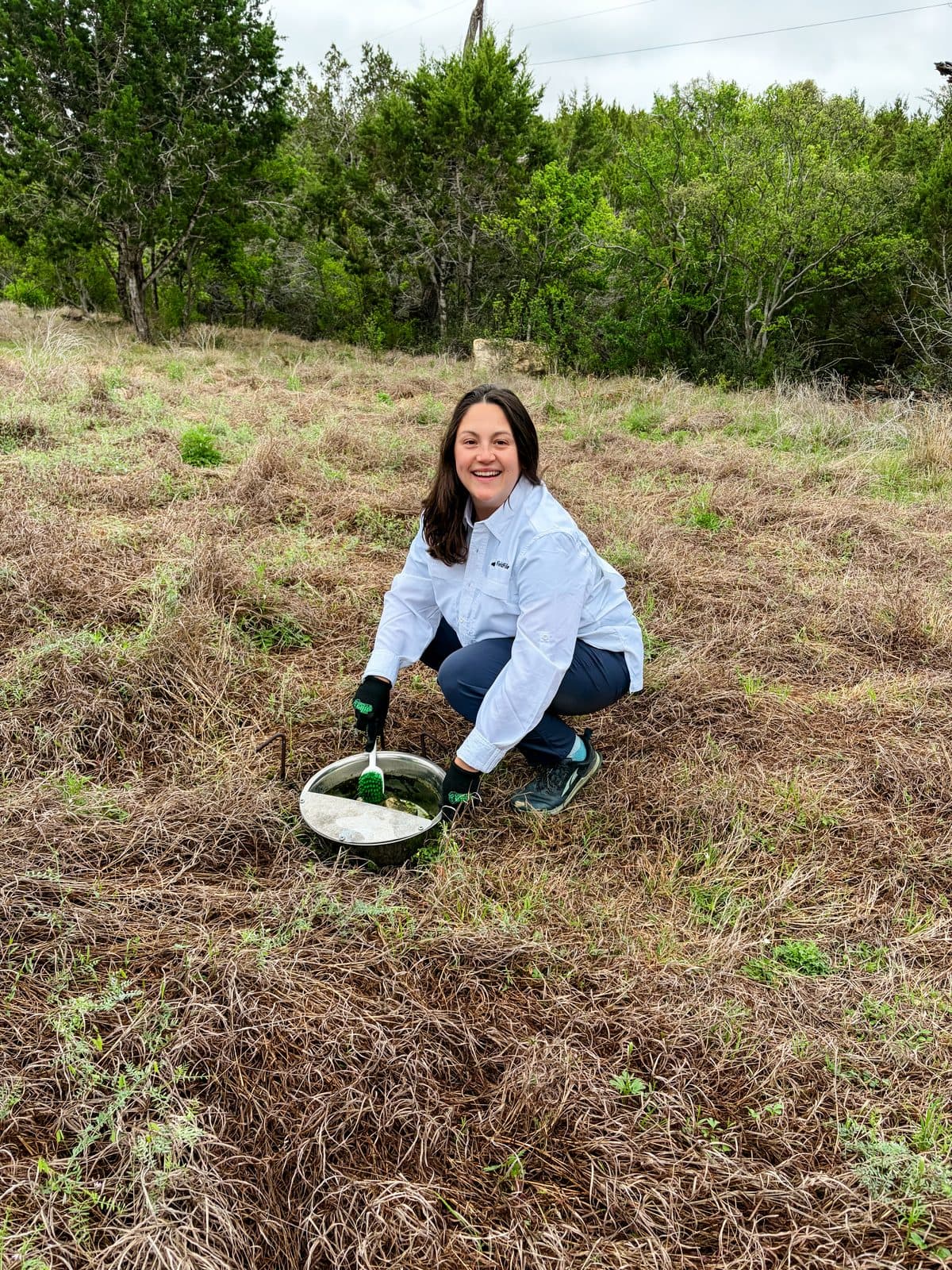 A landowner crouched in a field of dry grass, scrubbing algae out of the supplemental water bowl with a brush